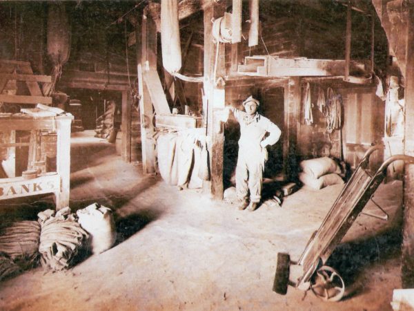 Floyd Powell stands in the Powell Feed Mill. The chute above his head was used for filling bags of grain, and the Fairbanks Scale on the left is designed specifically for weighing grain. Today the Powell Feed Mill is the commission sales barn in De Kalb Junction. Circa 1920s-1930s. De Kalb Junction, NY.