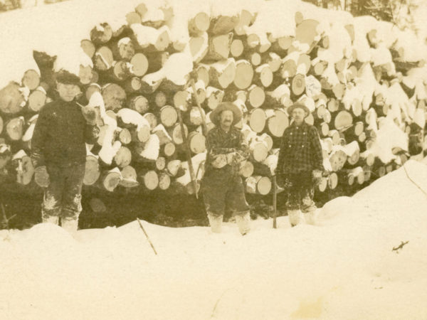 Loggers pose in front of snowcapped log pile in Long Lake