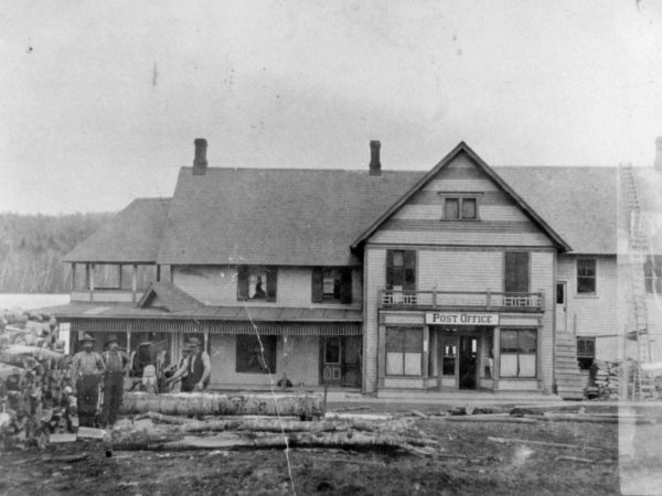 Men cutting firewood in front of a post office in Long Lake