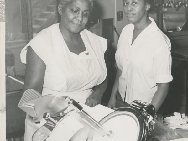 Two women with a meat slicer in the kitchen of the Echo Camp for Girls in Raquette Lake