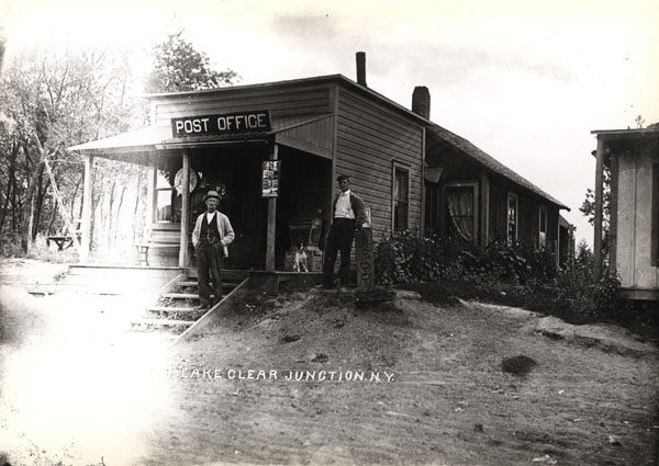 Two men and dog in front of post office in Lake Clear Junction