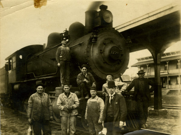 Eight men stand with locomotive at the junction in Lake Clear